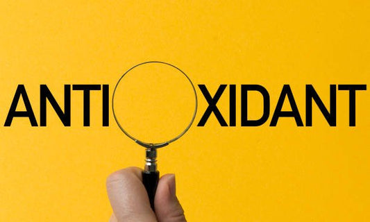 Hand holding a magnifying glass over the word 'ANTIOXIDANT' on a vibrant yellow background.