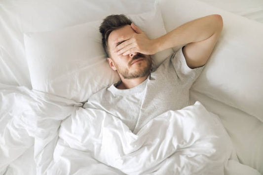 Man in bed covering face with hand, looking tired and stressed, surrounded by white bedding.