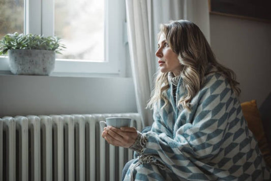 Woman wrapped in a blanket holding a cup, sitting by the window looking thoughtful and warm.