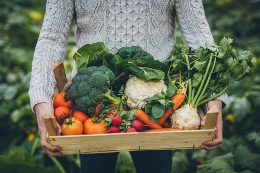 Person holding a wooden crate filled with fresh vegetables like broccoli, carrots, radishes, cauliflower, and tomatoes.