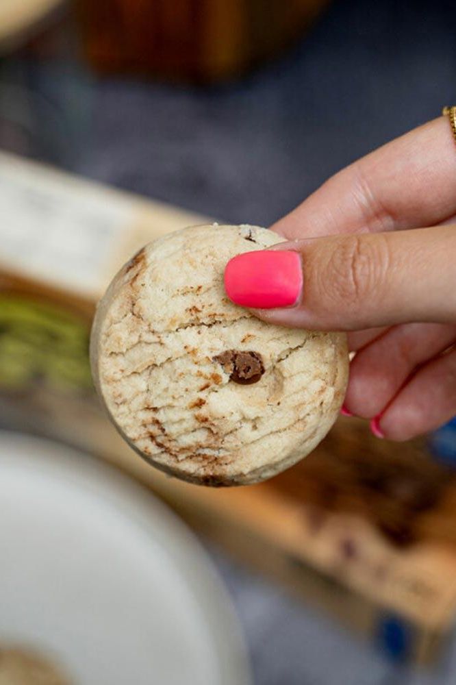 Hand holding a Nutriversum chocolate chip cookie with a pink nail polish, showcasing a healthy snack option.