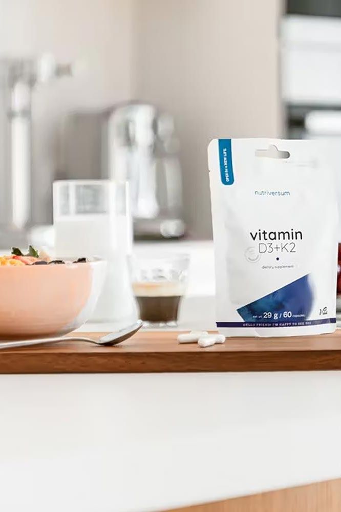 Nutriversum D3 + K2 vitamin capsules on a kitchen counter with healthy meal.