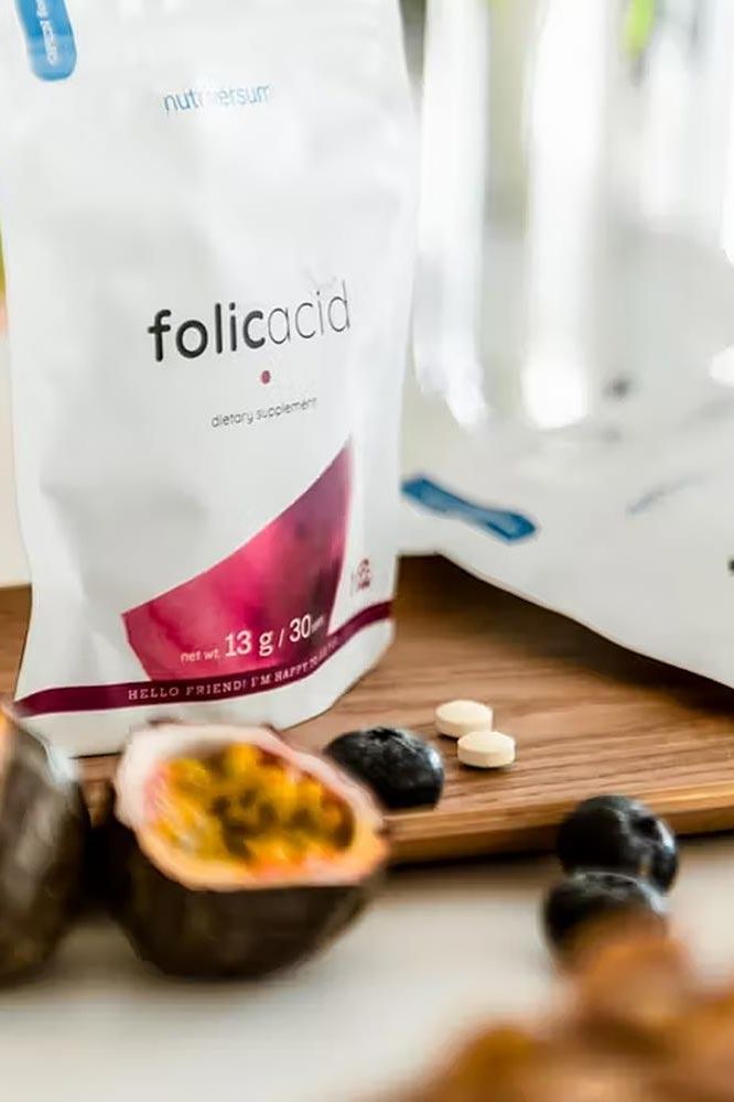 Nutriversum Folic Acid supplement packaging with tablets on wooden surface and fruits in the background.