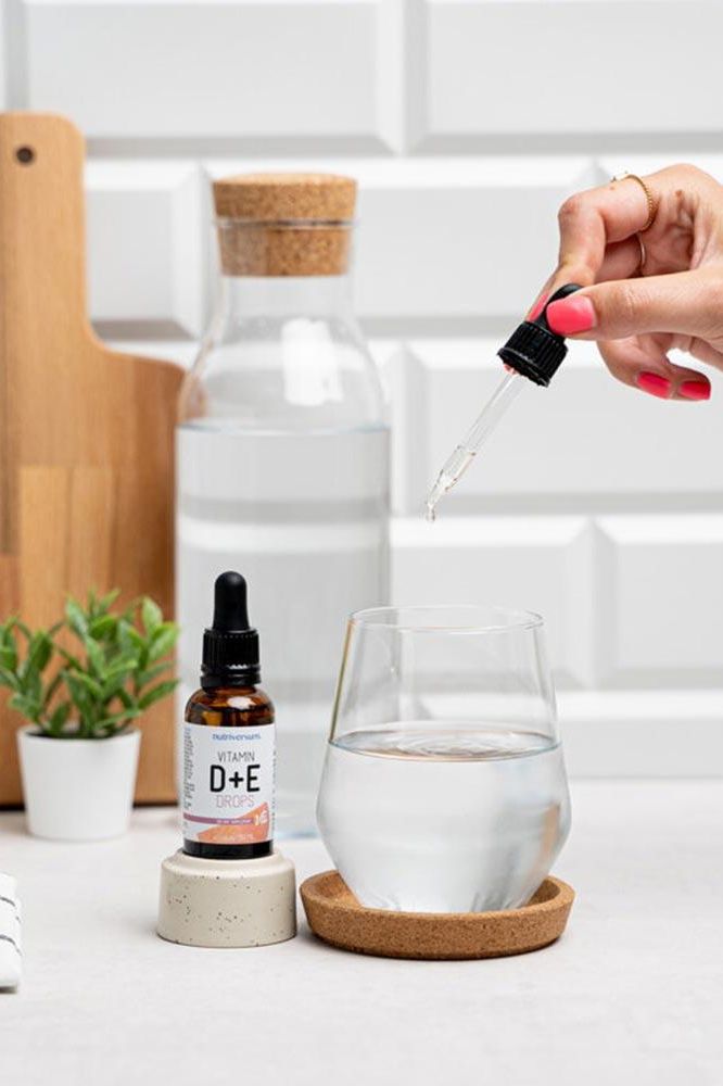 Hand adding Nutriversum Vitamin D+E drops to a glass of water on a kitchen counter.