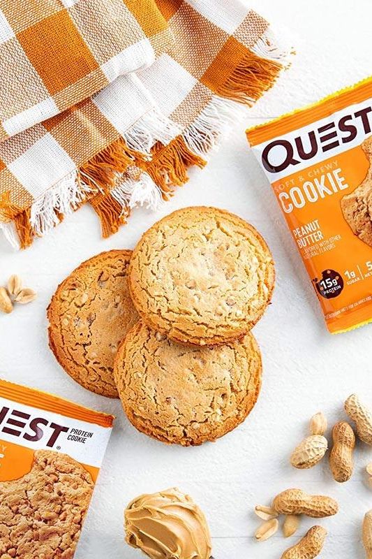 Quest Nutrition Protein Cookie with peanut butter, beside unpackaged cookies and peanuts on a checkered cloth.