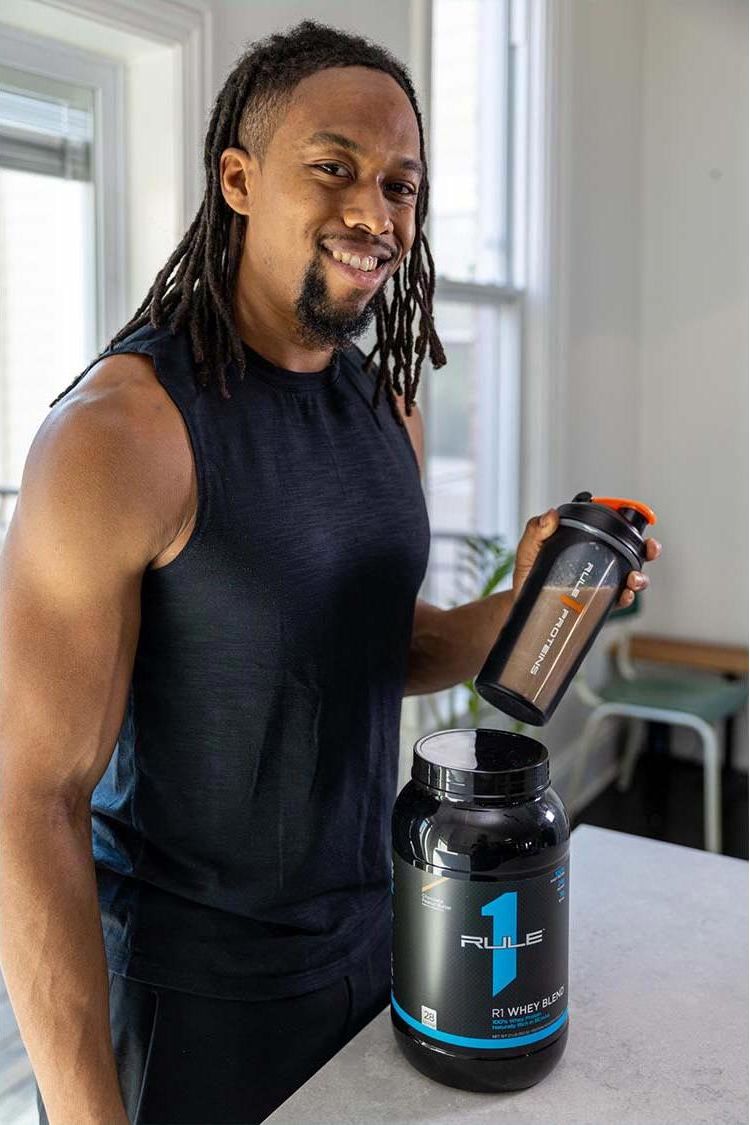Smiling man holding Rule One R1 Whey Blend protein powder and shaker in kitchen, showcasing fitness lifestyle.