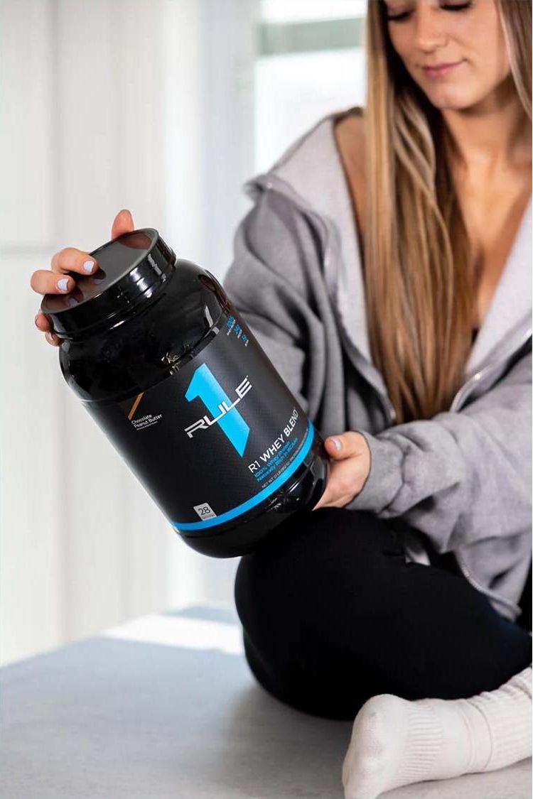 Woman holding Rule One R1 Whey Blend protein tub, showcasing the product's branding and packaging.