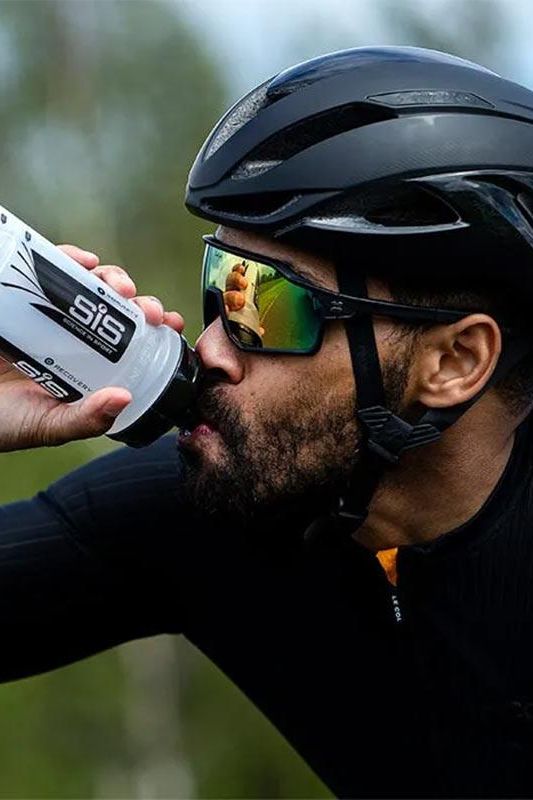 Athlete drinking from a Science in Sport water bottle while cycling, wearing a helmet and sunglasses.
