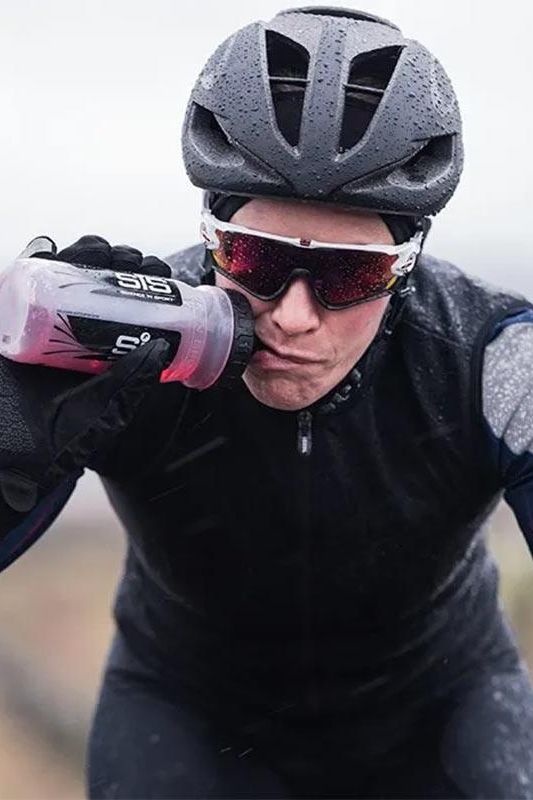 Cyclist drinking from a Science in Sport water bottle during rainy weather, showcasing hydration for optimal performance.