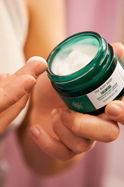 Person holding The Body Shop Smoothing Day Cream jar with cream on finger.