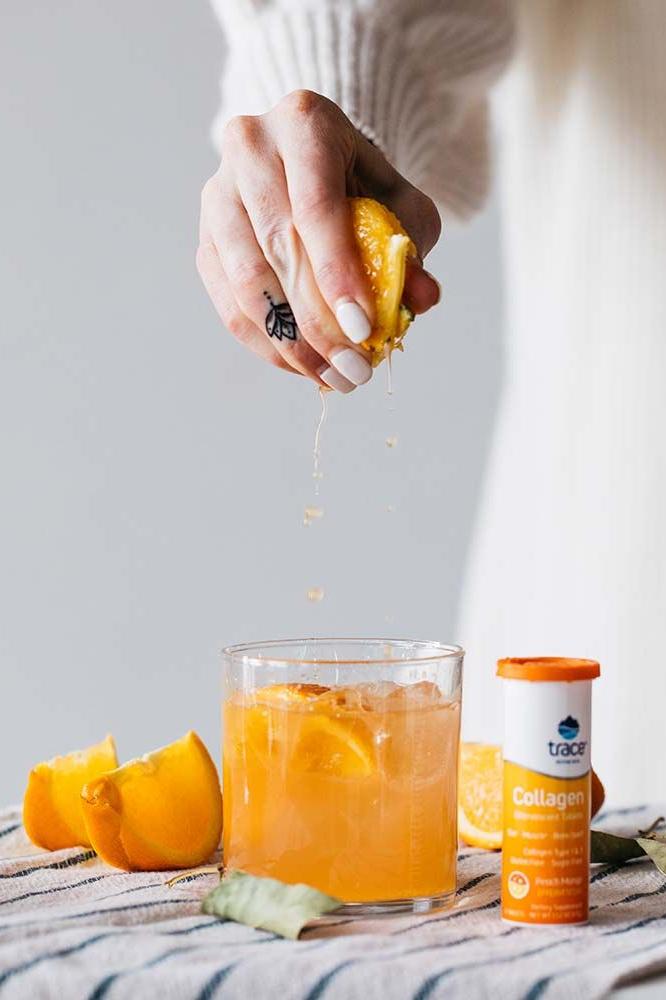 Person squeezing fresh peach over a glass of peach mango collagen drink with Trace Minerals tablet.