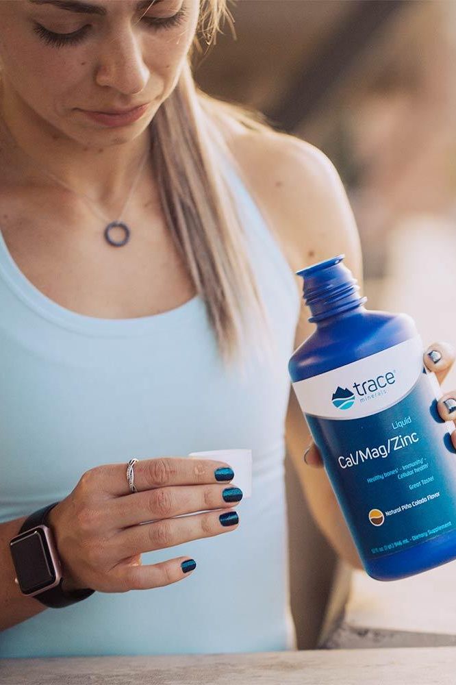 Woman holding Trace Minerals Liquid Cal/Mag/Zinc bottle with Piña Colada flavor in a fitness setting.