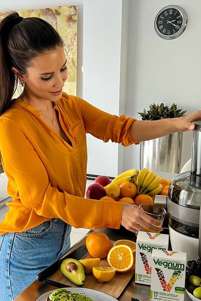 A woman in a yellow shirt preparing juice with fresh fruits and Vegnum - Multi-D Plus capsules on the counter.