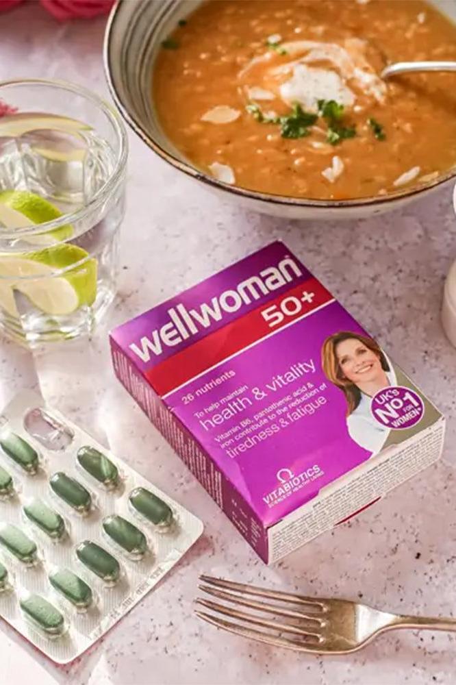 Wellwoman 50+ tablets beside a bowl of soup and a glass of water with lime, promoting health for women over 50.